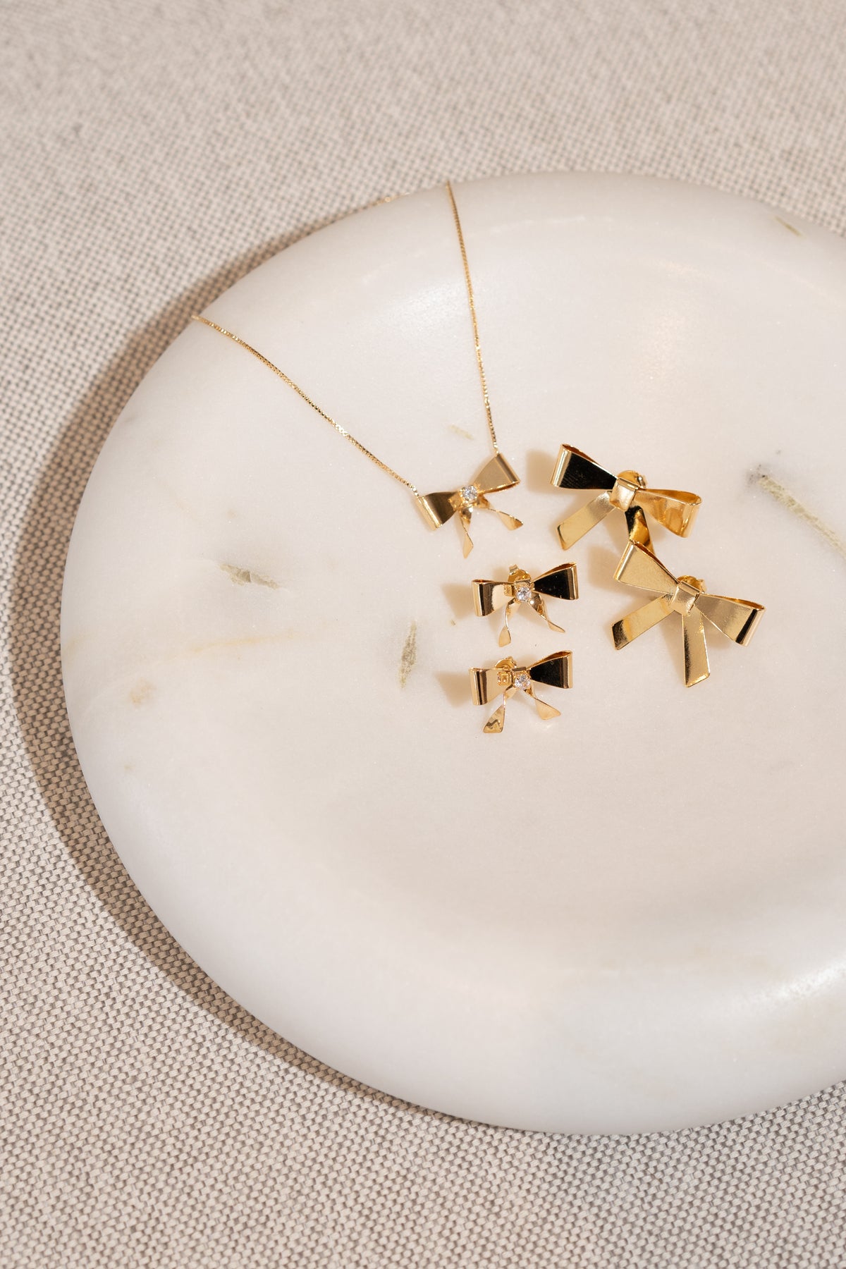 A matching gold jewelry set featuring bow-shaped pendant necklaces and earrings rests elegantly on a white marble tray.