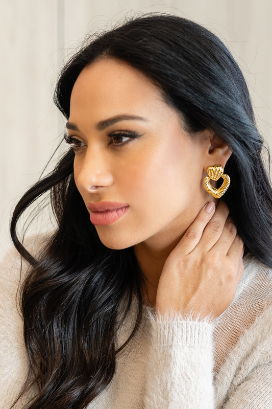 A dark-haired woman wearing textured gold heart-shaped statement earrings gently touches her neck.