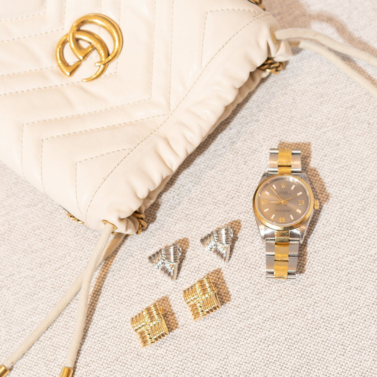 A cream-colored Gucci quilted leather handbag rests beside a two-tone Rolex wristwatch and pairs of gold and silver statement earrings on a neutral textured surface.