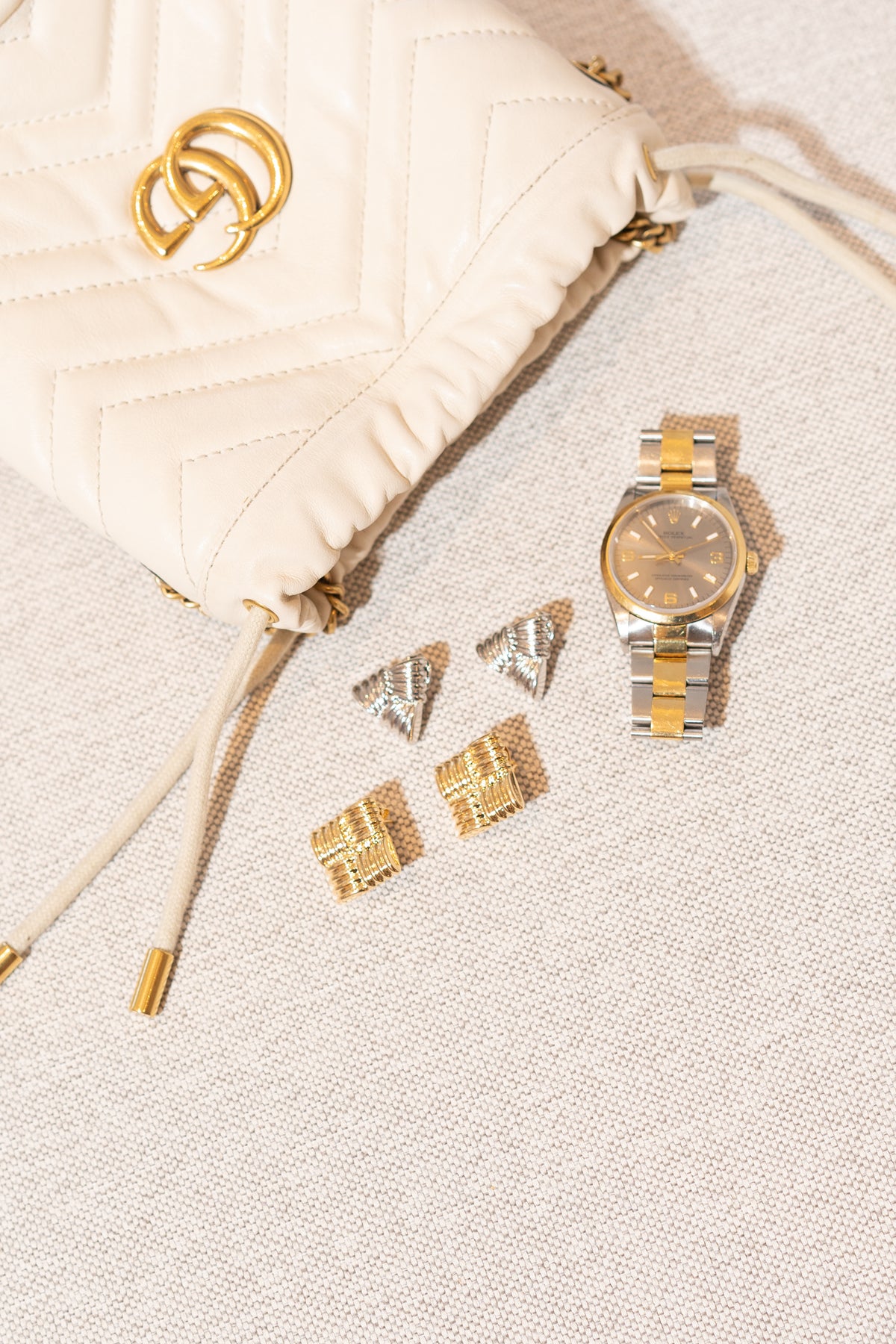 A cream quilted Gucci handbag rests beside a two-tone Rolex watch and two pairs of designer earrings on a textured surface.