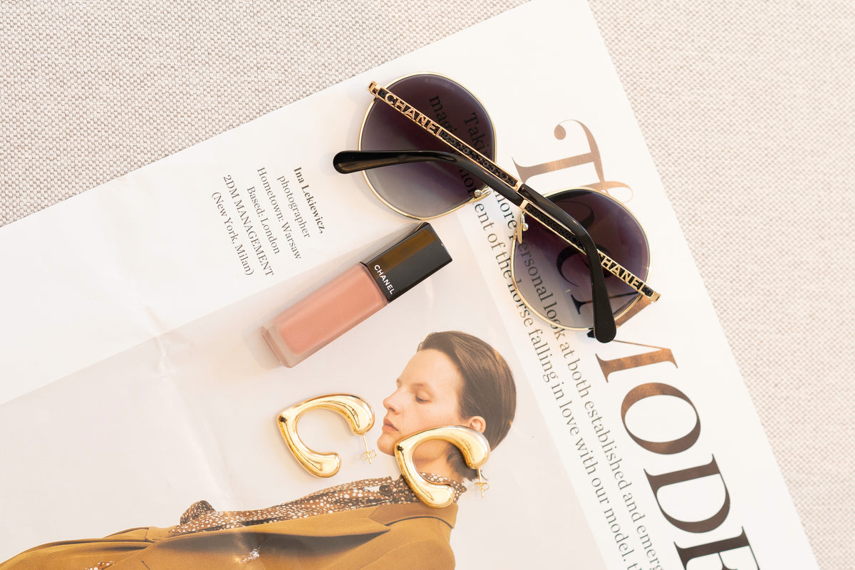 Chanel sunglasses, a Chanel lipstick, and gold hoop earrings rest on an open magazine page.
