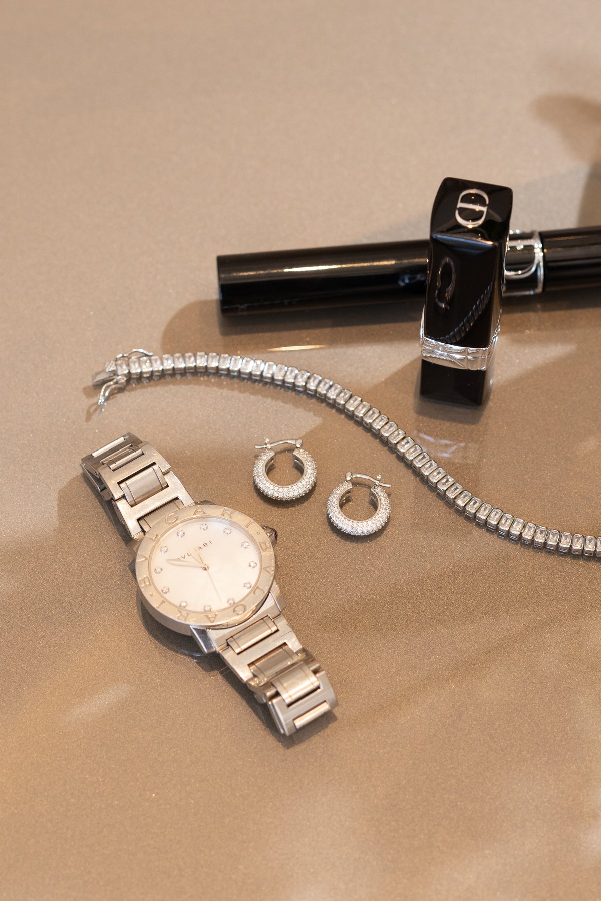 A silver Bvlgari wristwatch, diamond hoop earrings, a sparkling tennis bracelet, and a black Dior lipstick tube rest together on a reflective surface.