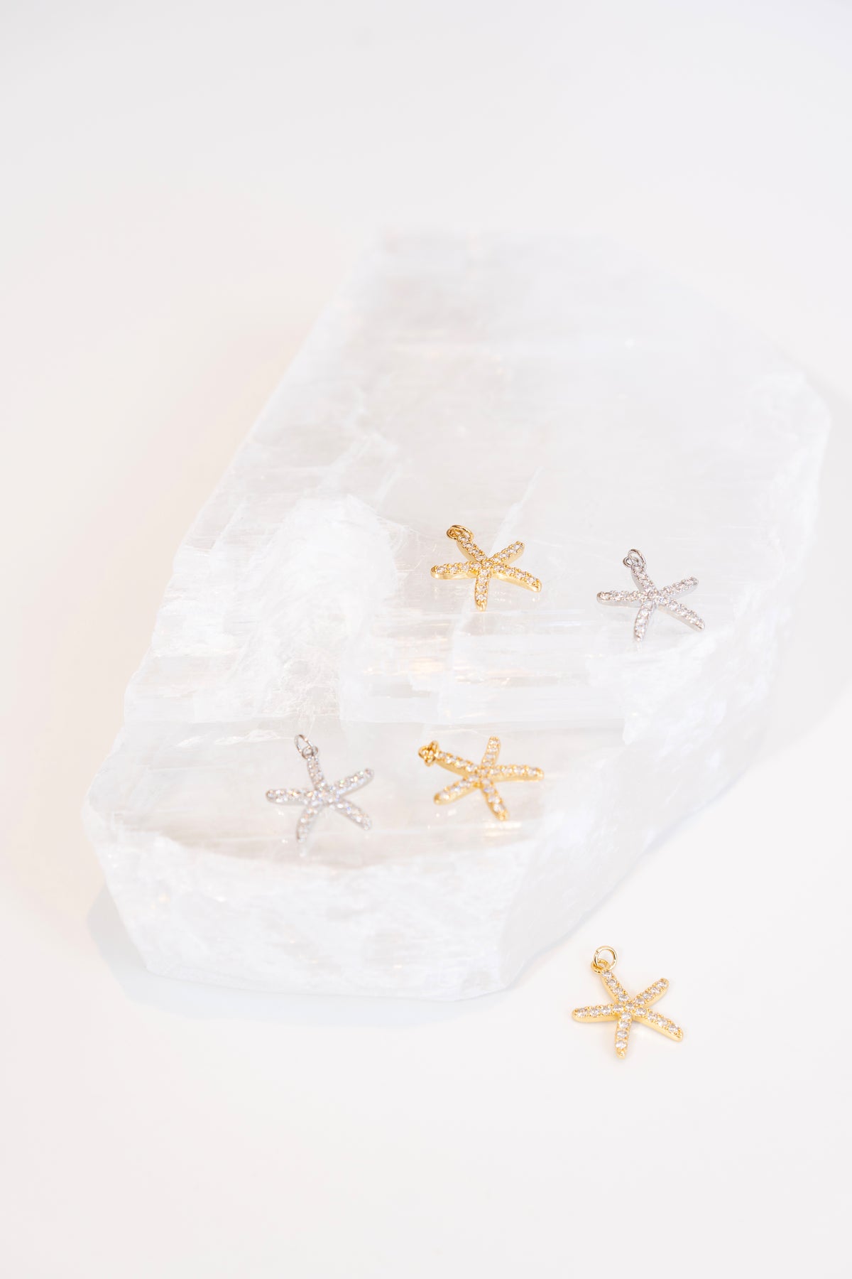Sparkling pave-set gold and silver starfish charms rest delicately on a white crystal display block.
