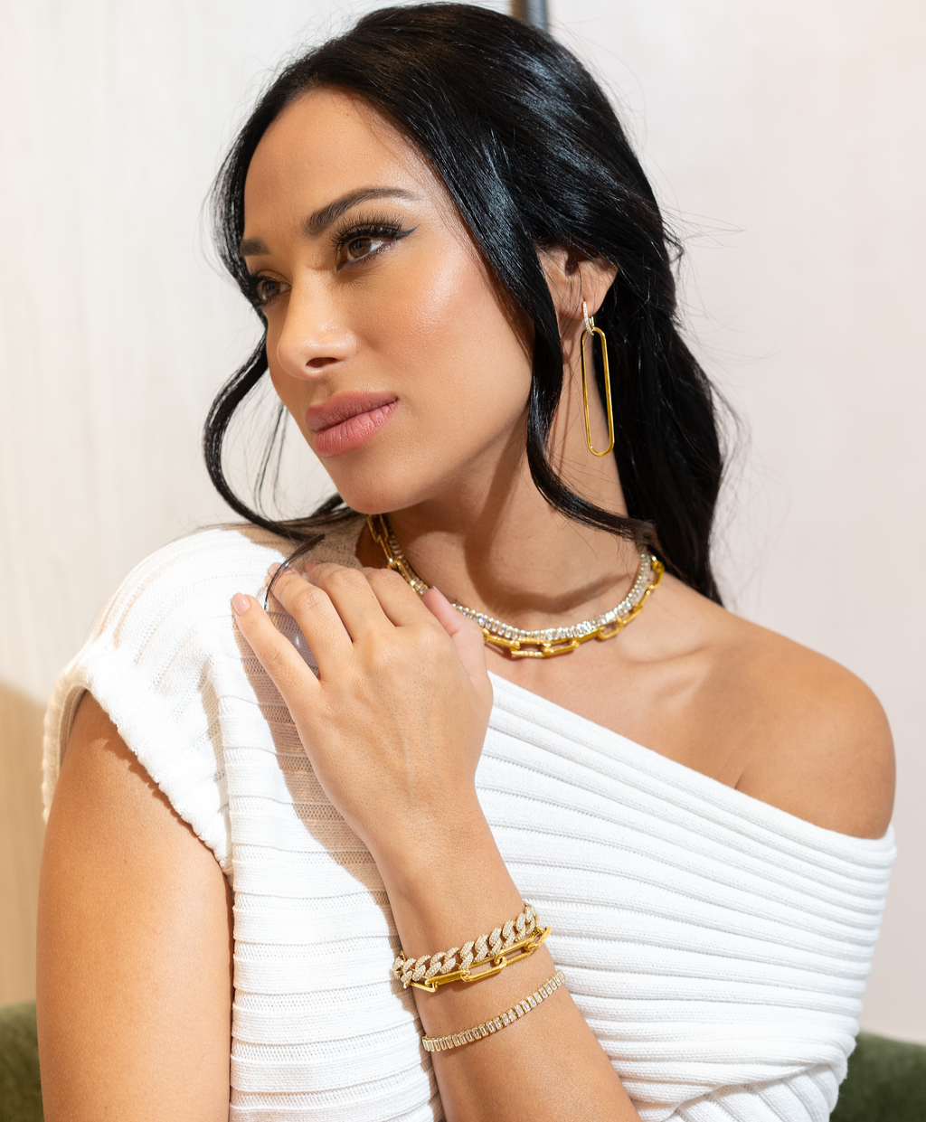 A stylish woman wearing a white off-the-shoulder sweater showcases gold jewelry, including layered necklaces, chunky chain bracelets, and geometric earrings.
