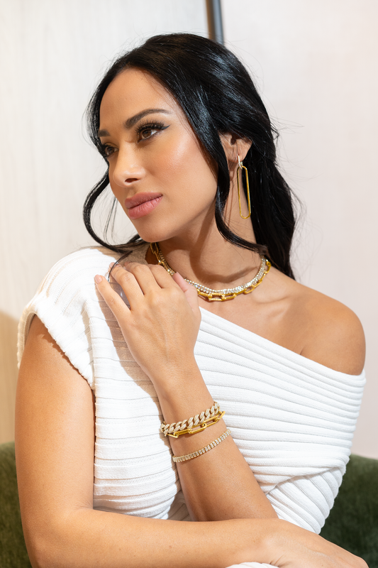 A dark-haired woman wearing a white off-the-shoulder sweater showcases layered gold jewelry, including a chain necklace, matching bracelets, and geometric earrings.