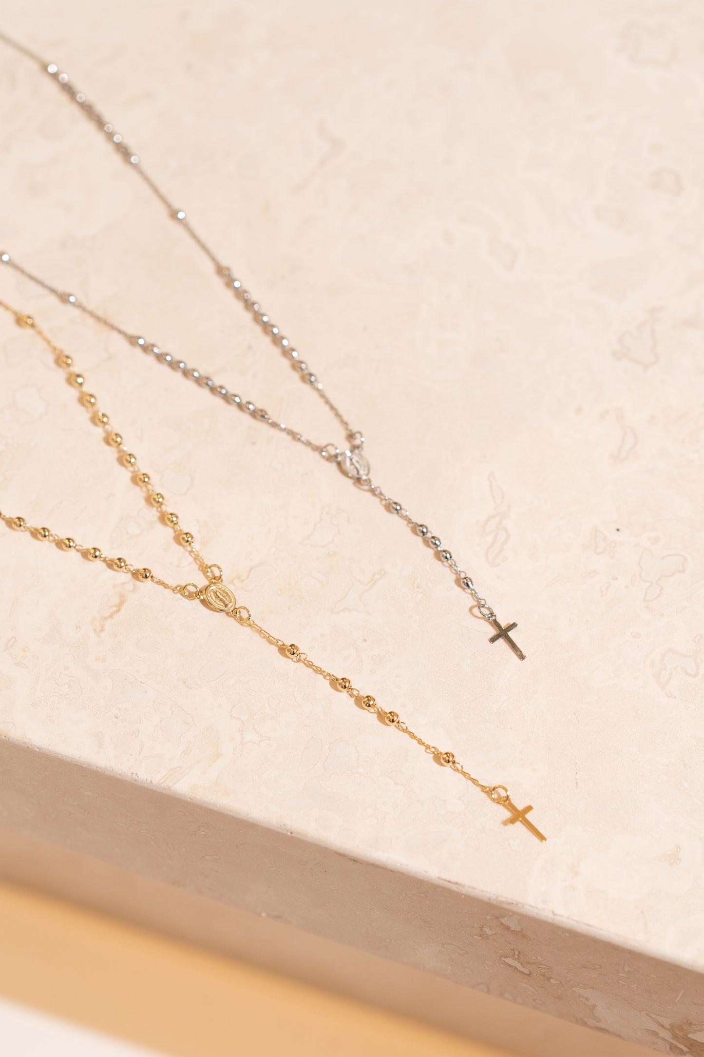 A gold rosary necklace and a silver rosary necklace rest elegantly on a textured beige surface.