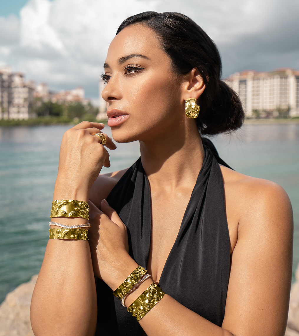 A stylish woman wearing a black halter top showcases chunky gold textured cuffs and matching earrings by the water.