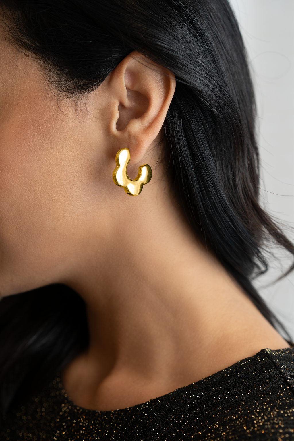 A woman with dark hair wears a unique, chunky gold abstract-shaped hoop earring on her earlobe.