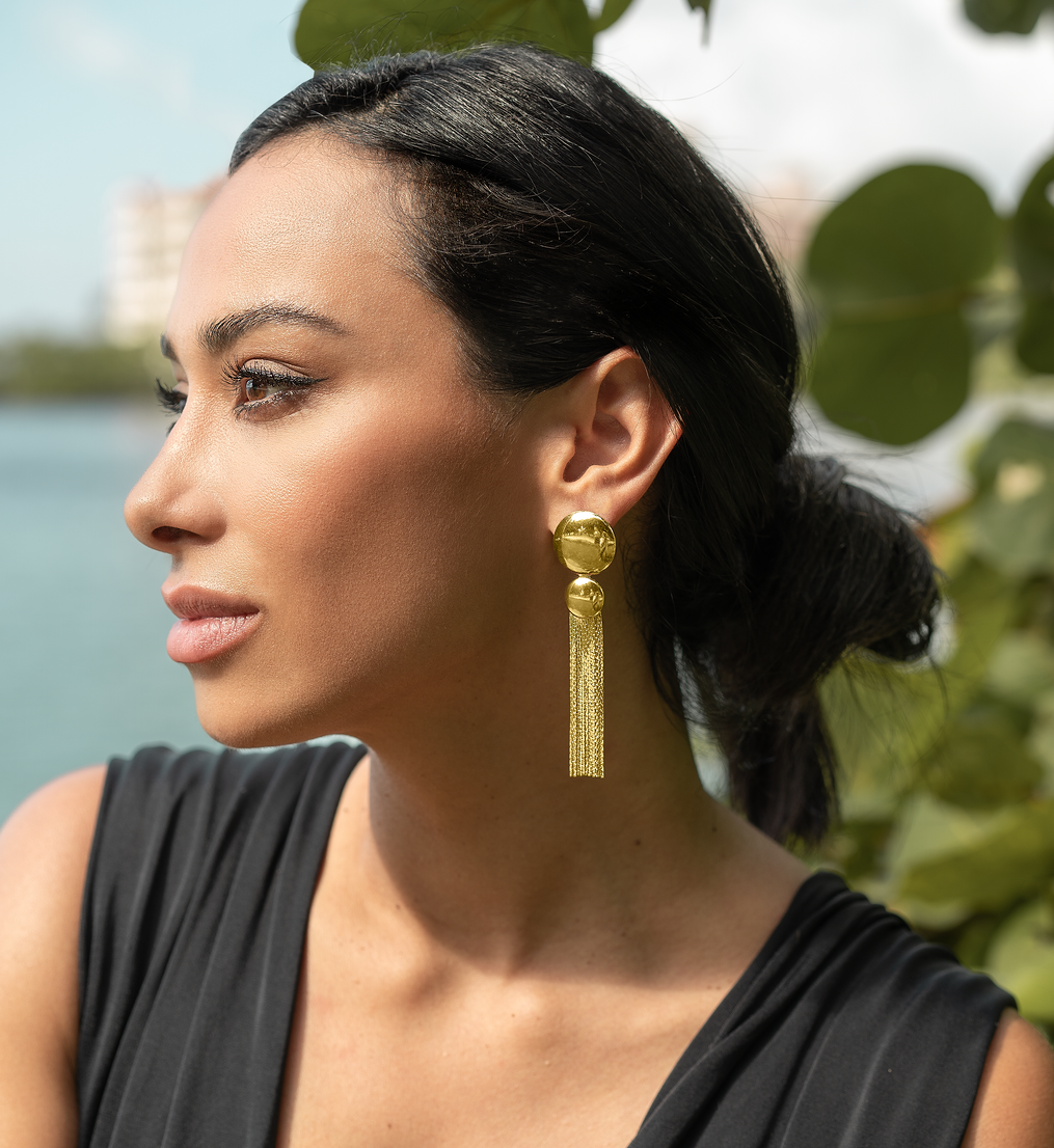 A woman with dark hair wearing statement gold fringe earrings poses outdoors near the water.