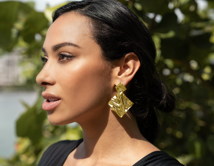 A woman with dark hair wears bold, gold statement earrings outdoors against a leafy green background.