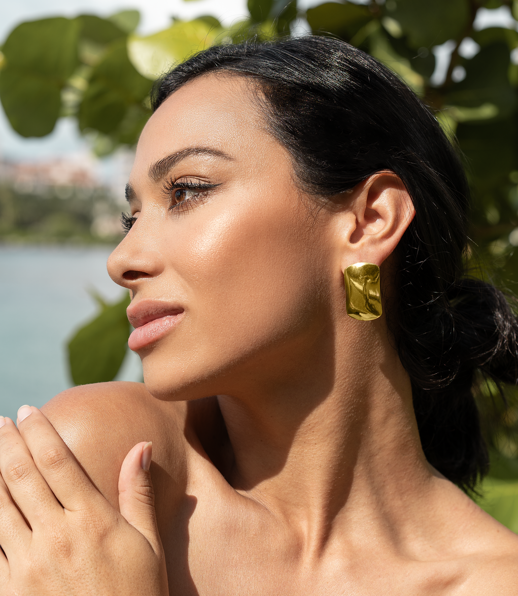 A dark-haired woman wearing bold gold statement earrings poses outdoors near lush green foliage.