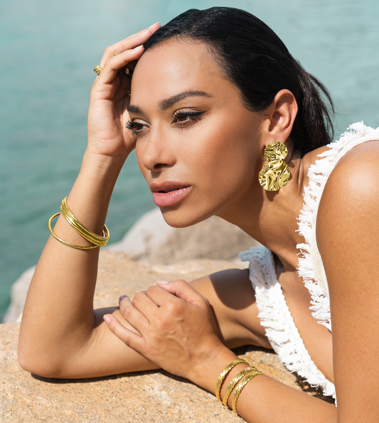 A stylish woman wearing statement gold earrings and stacked bracelets poses gracefully against a backdrop of water and rocks.
