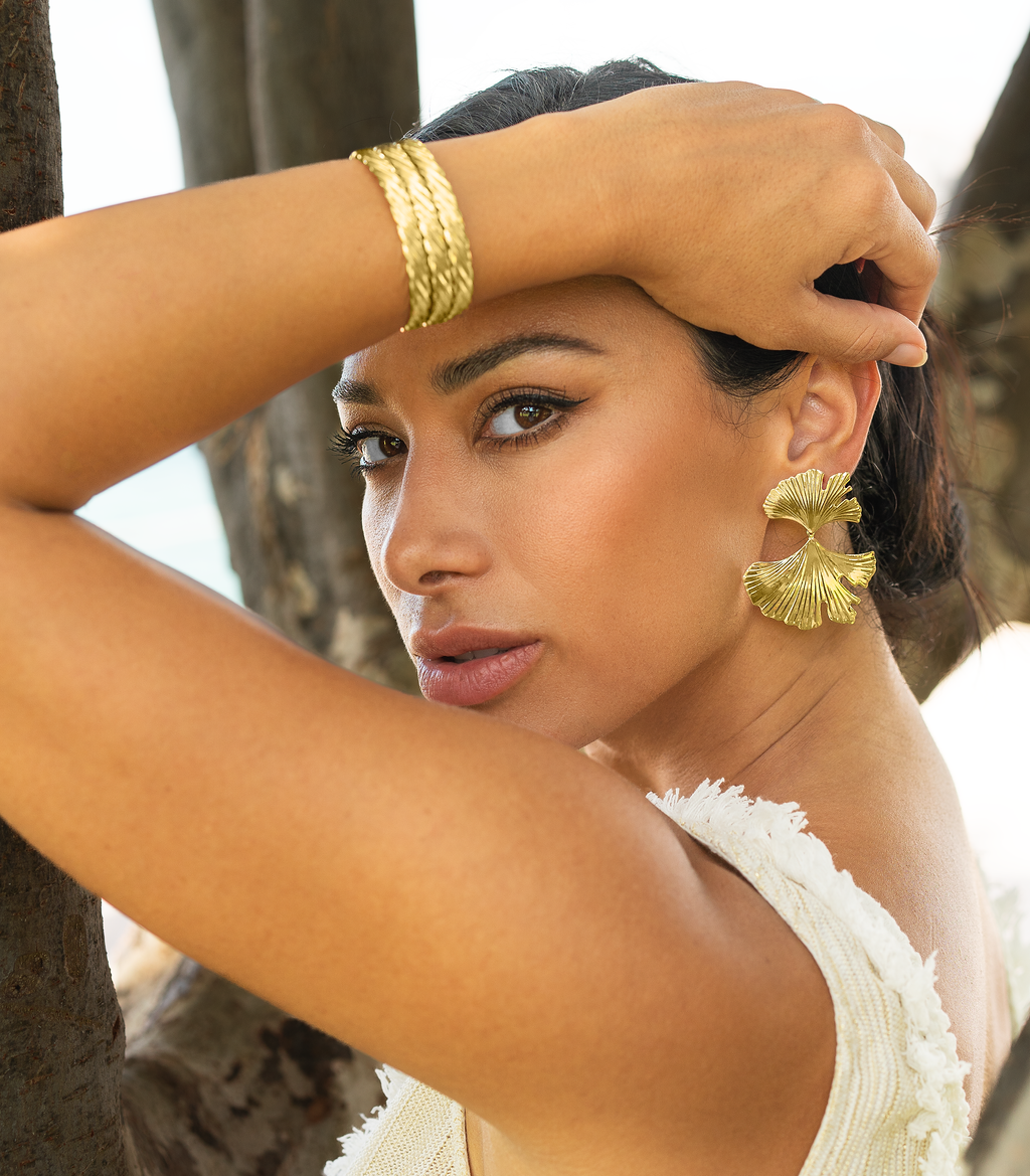 A stylish woman wearing statement gold ginkgo leaf earrings and a braided cuff bracelet gently poses against a tree trunk.