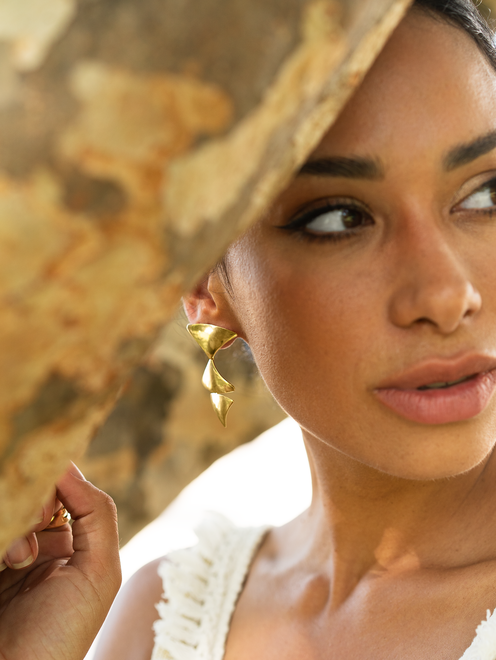 A young woman poses outdoors, wearing bold gold statement earrings with geometric triangular shapes while partially framed by a rustic wooden branch.
