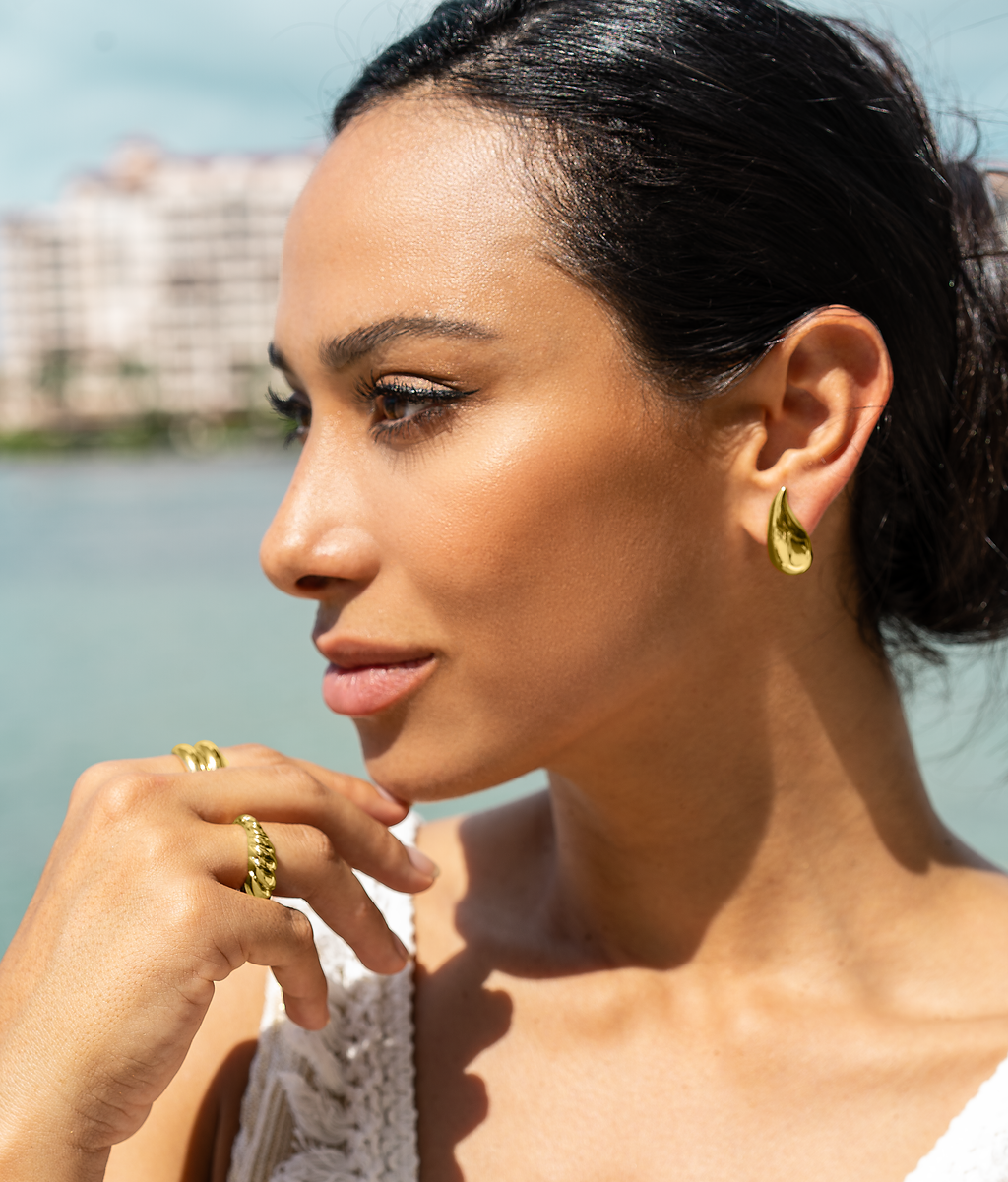 A stylish woman with dark hair wears teardrop gold earrings and statement gold rings while posing outdoors near the water.
