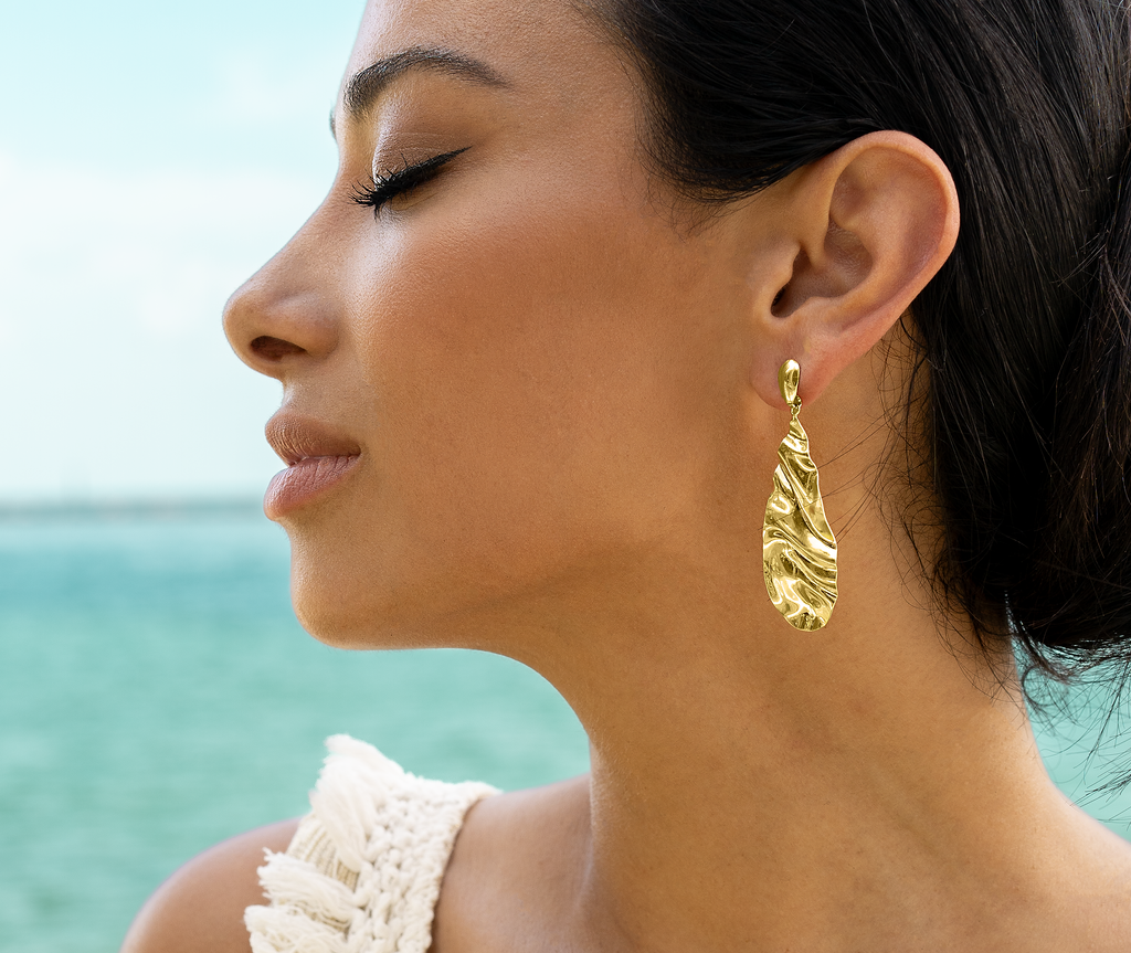 A woman in profile showcases a textured, teardrop-shaped gold dangle earring against an ocean backdrop.