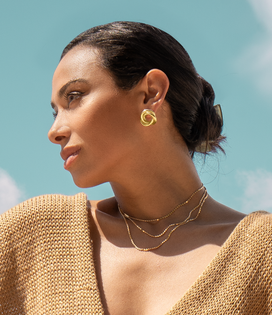 A woman with sleek dark hair styled in a bun wears chunky gold knot earrings and layered gold beaded necklaces against a bright blue sky.