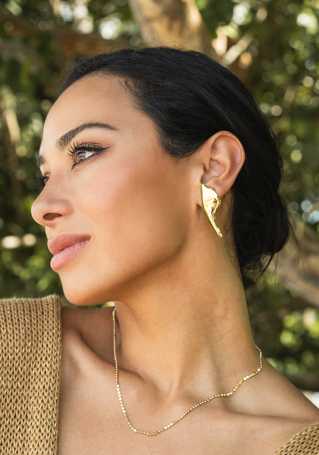 A woman with dark hair wears a striking gold statement earring and a delicate matching chain necklace.