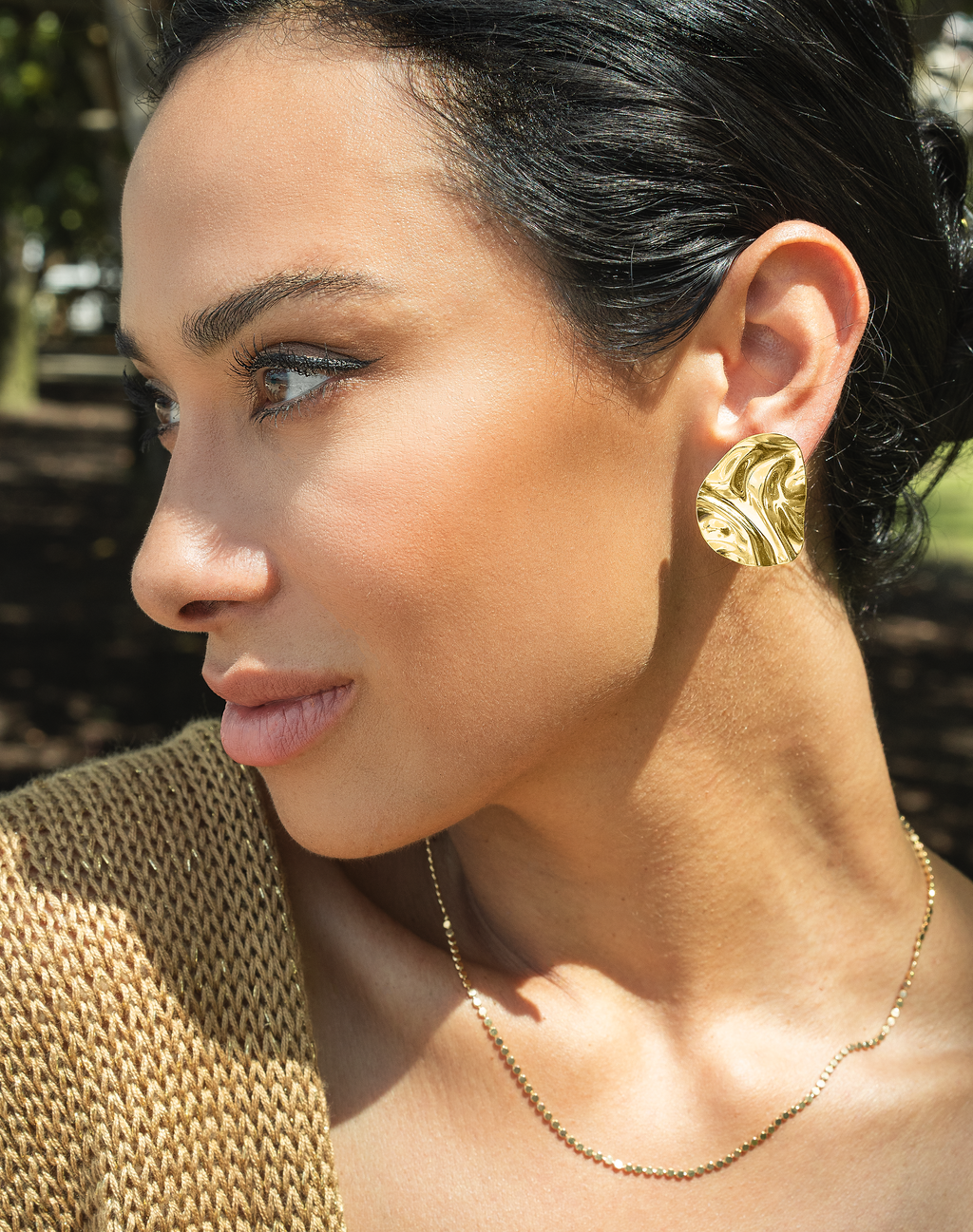 A stylish woman with dark hair wears a large, hammered gold statement earring and a delicate chain necklace outdoors.