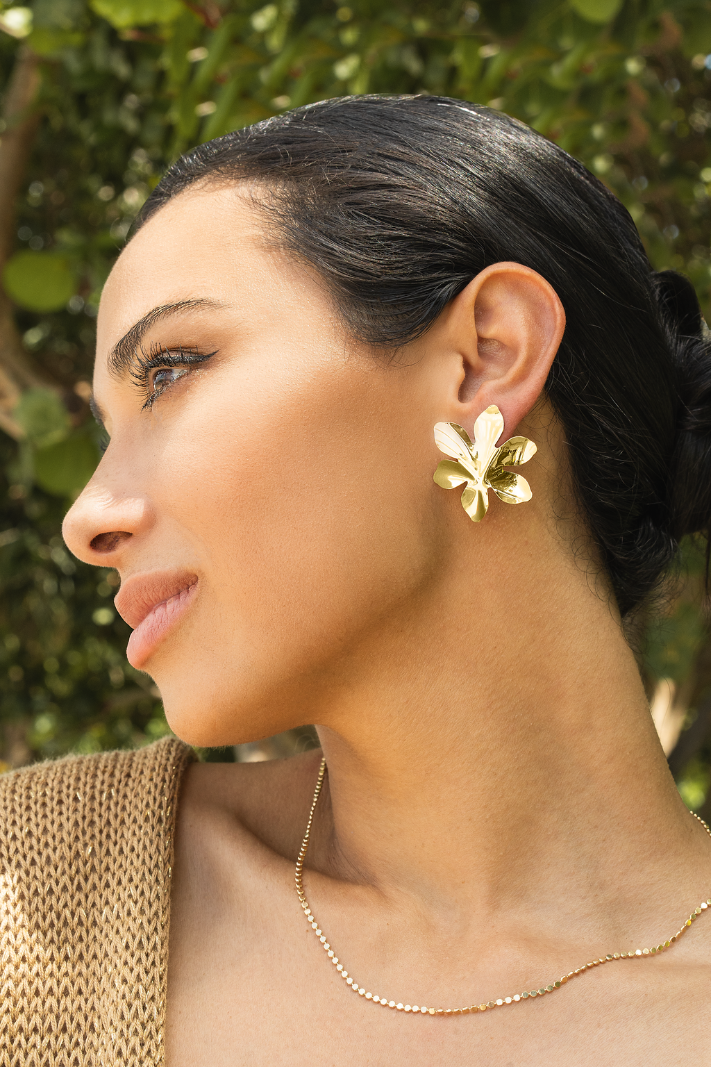 A dark-haired woman showcases polished gold floral statement earrings outdoors against a leafy green backdrop.