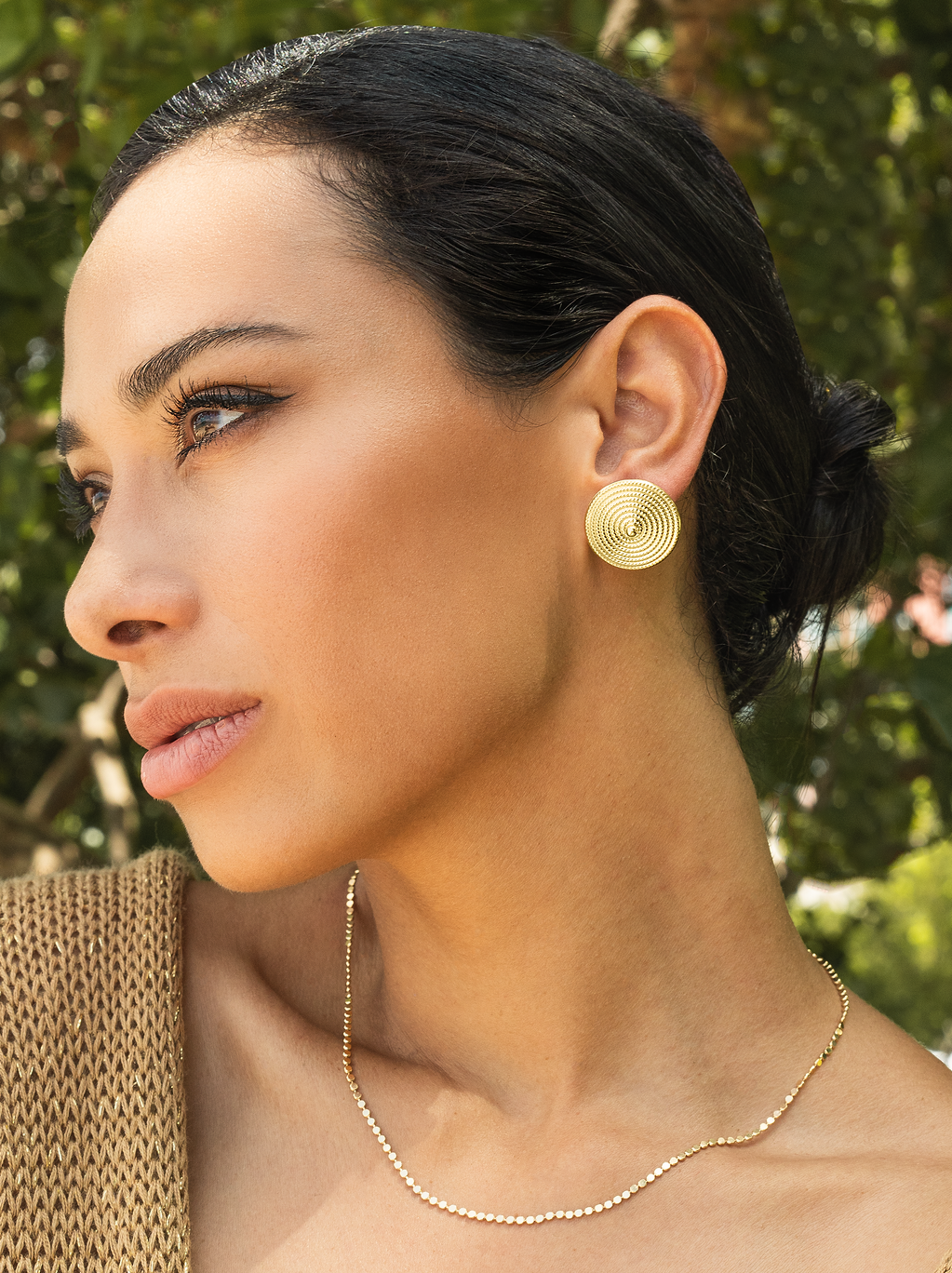 A woman with dark hair in a bun wears large, textured gold circular stud earrings outdoors.