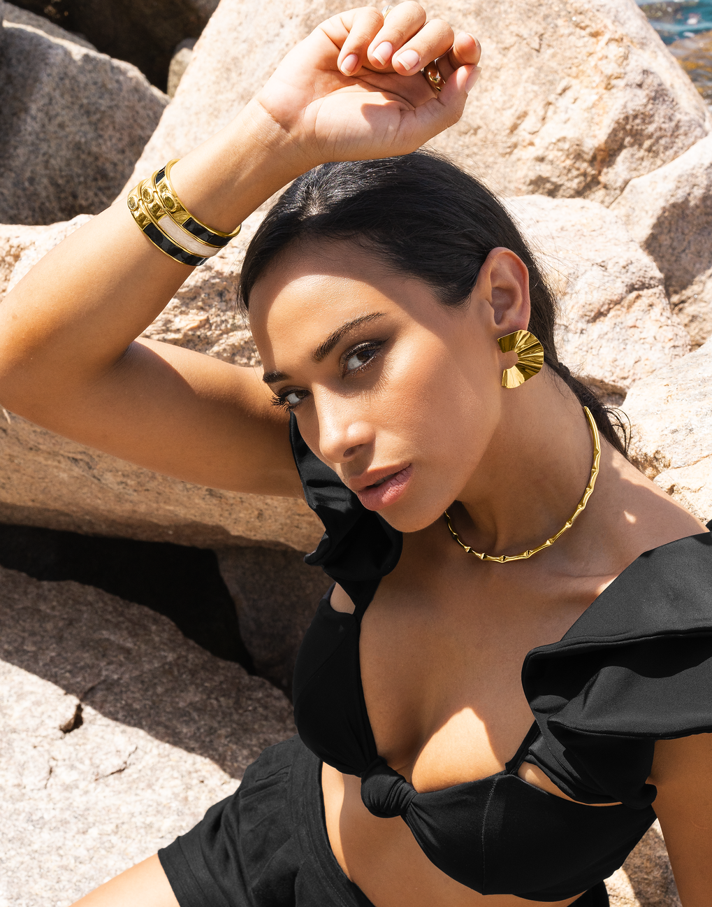 A dark-haired woman in a black bikini poses among sunlit rocks while wearing bold gold jewelry, including textured fan earrings, a bamboo-style choker necklace, and matching enamel bangles on her raised arm.