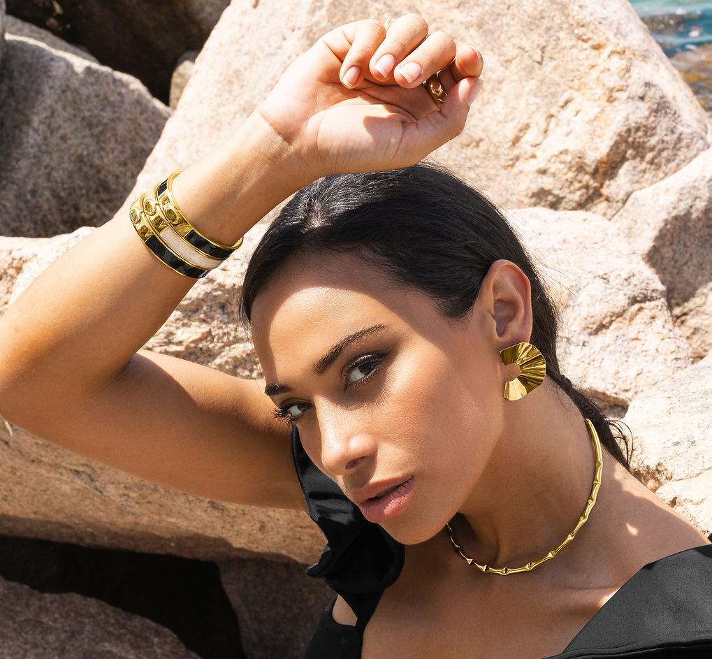 A glamorous woman wearing statement gold designer jewelry poses against a rocky coastal backdrop.