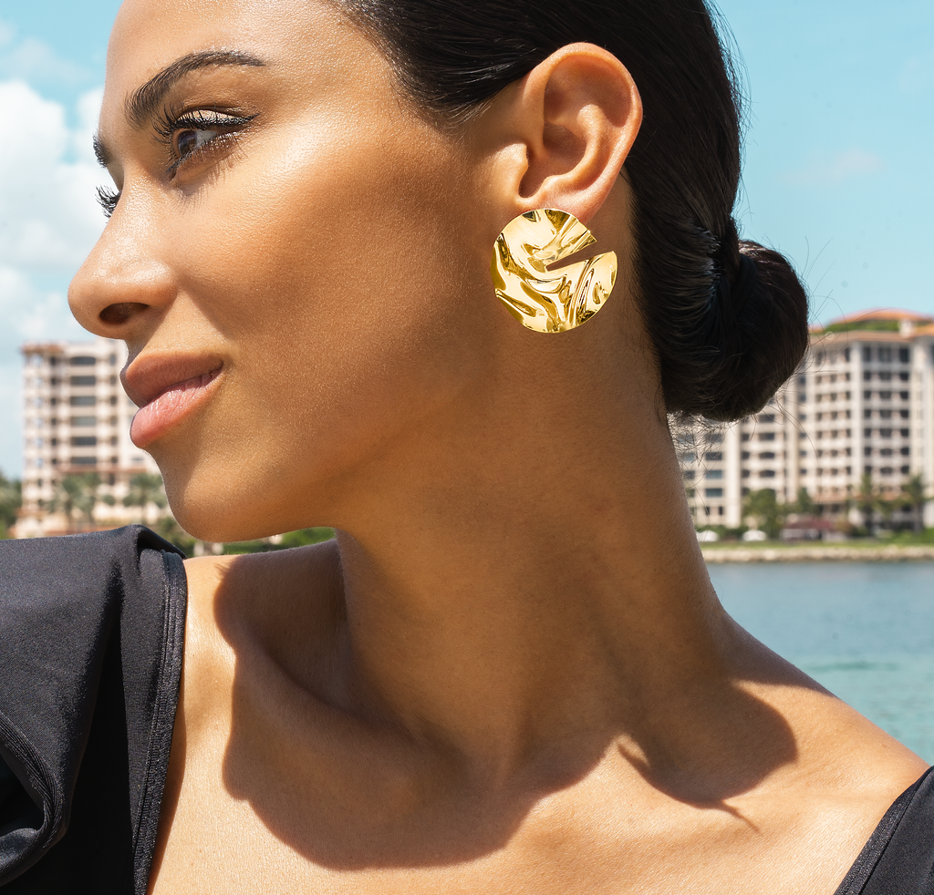 A glamorous woman wearing a large, textured gold statement earring poses outdoors by the water.