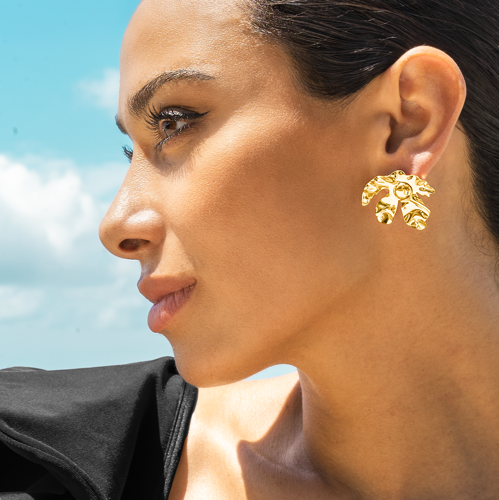 A woman wearing a statement gold flower earring poses outdoors against a bright blue sky.