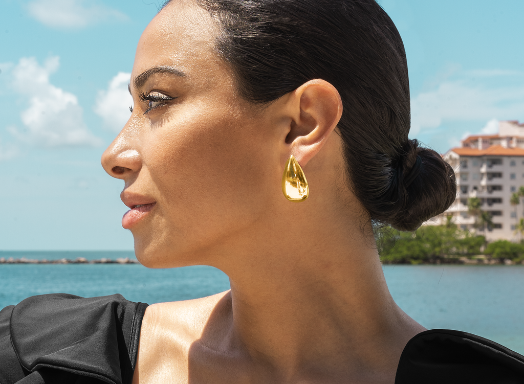 A woman with dark hair in a bun wears a shiny gold teardrop earring by the water.