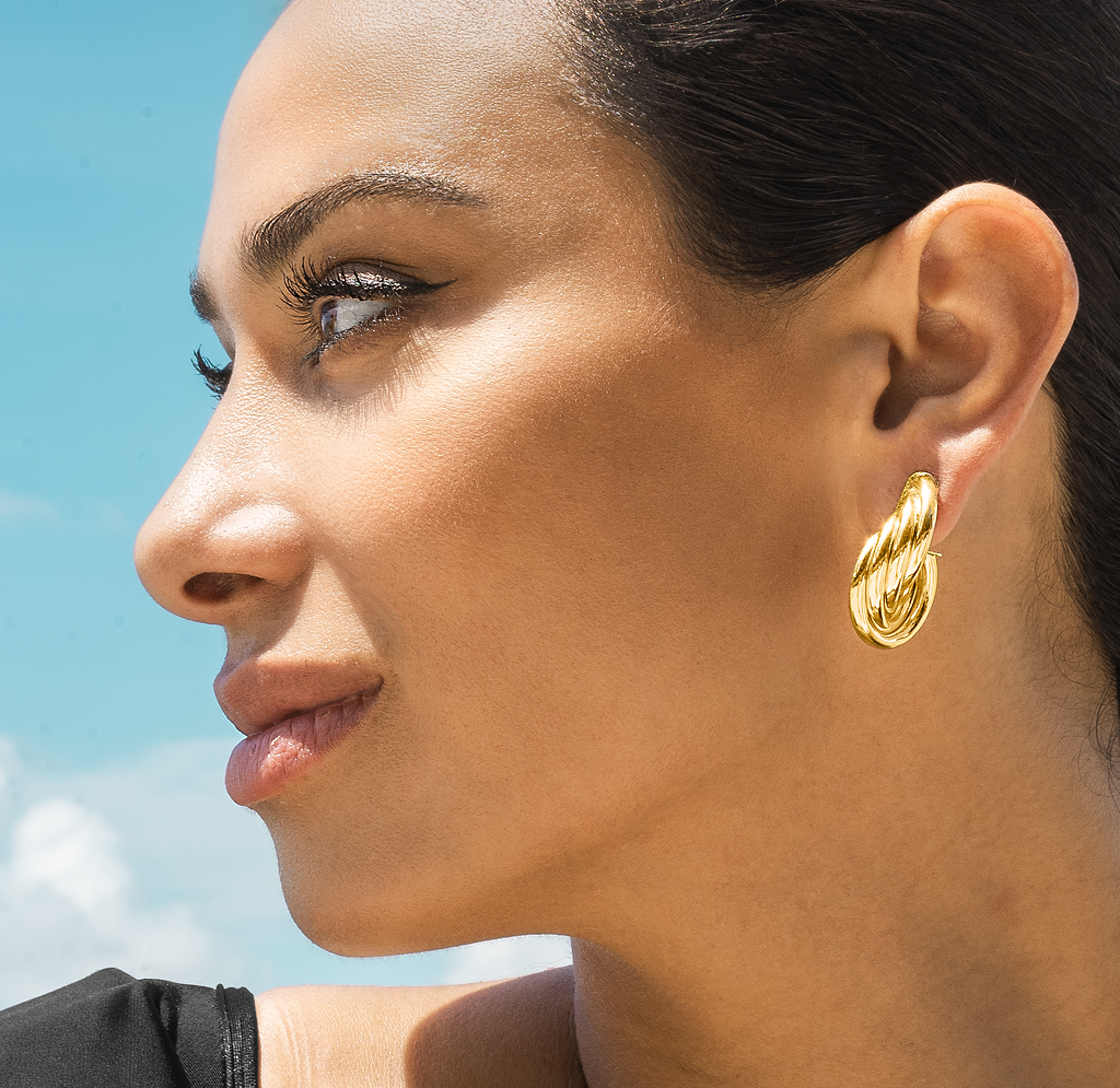 A woman with dark hair in profile wears bold gold triple-hoop earrings against a bright blue sky.