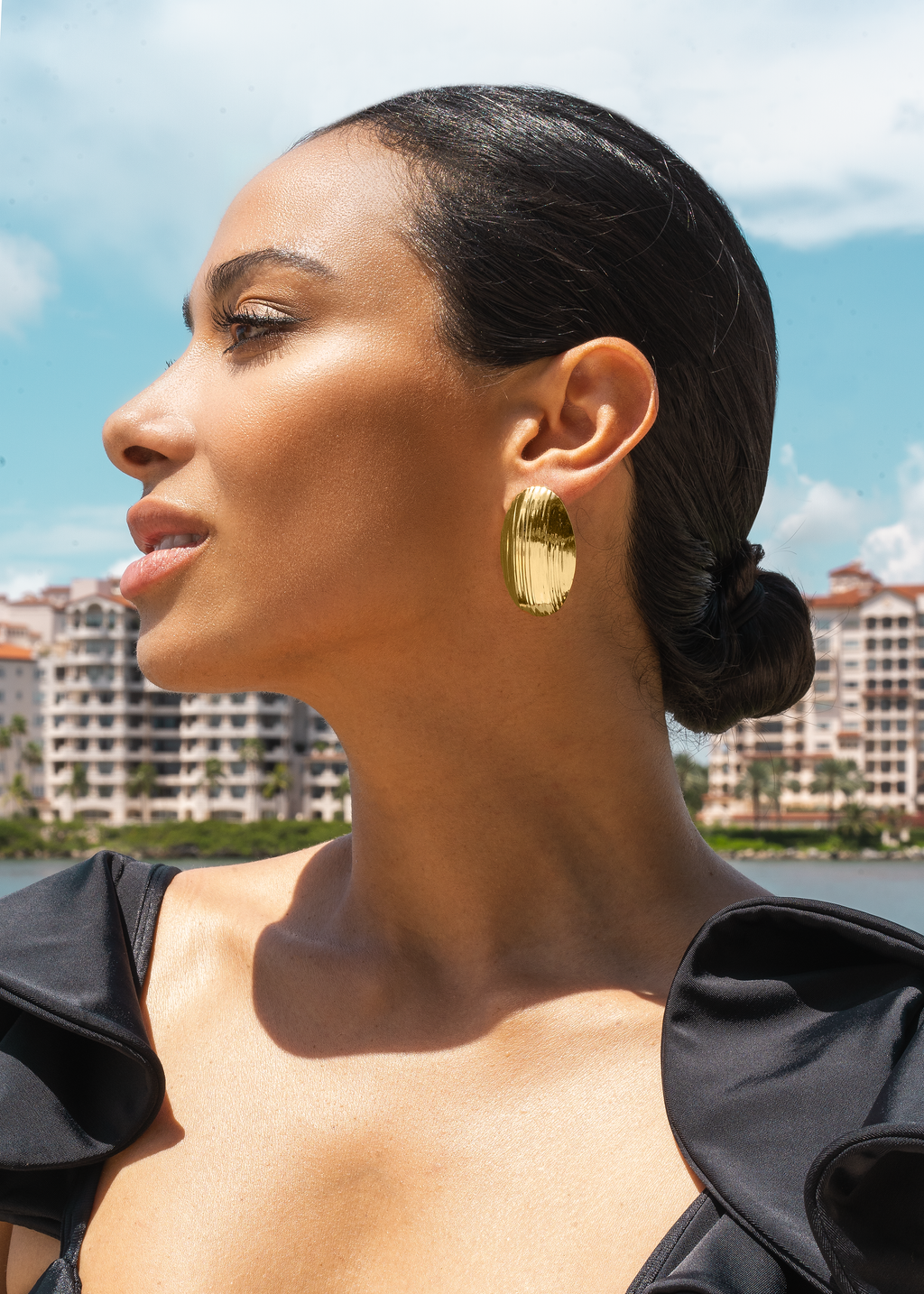 A profile view of a woman wearing striking gold textured oval statement earrings against a sunny waterfront backdrop.