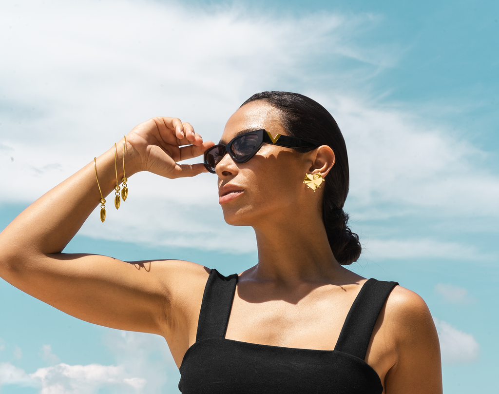 A stylish woman wearing chic black sunglasses, statement gold heart earrings, and layered gold bracelets poses outdoors against a bright blue sky.