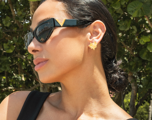 A stylish woman wearing black cat-eye sunglasses showcases a textured gold heart stud earring outdoors.