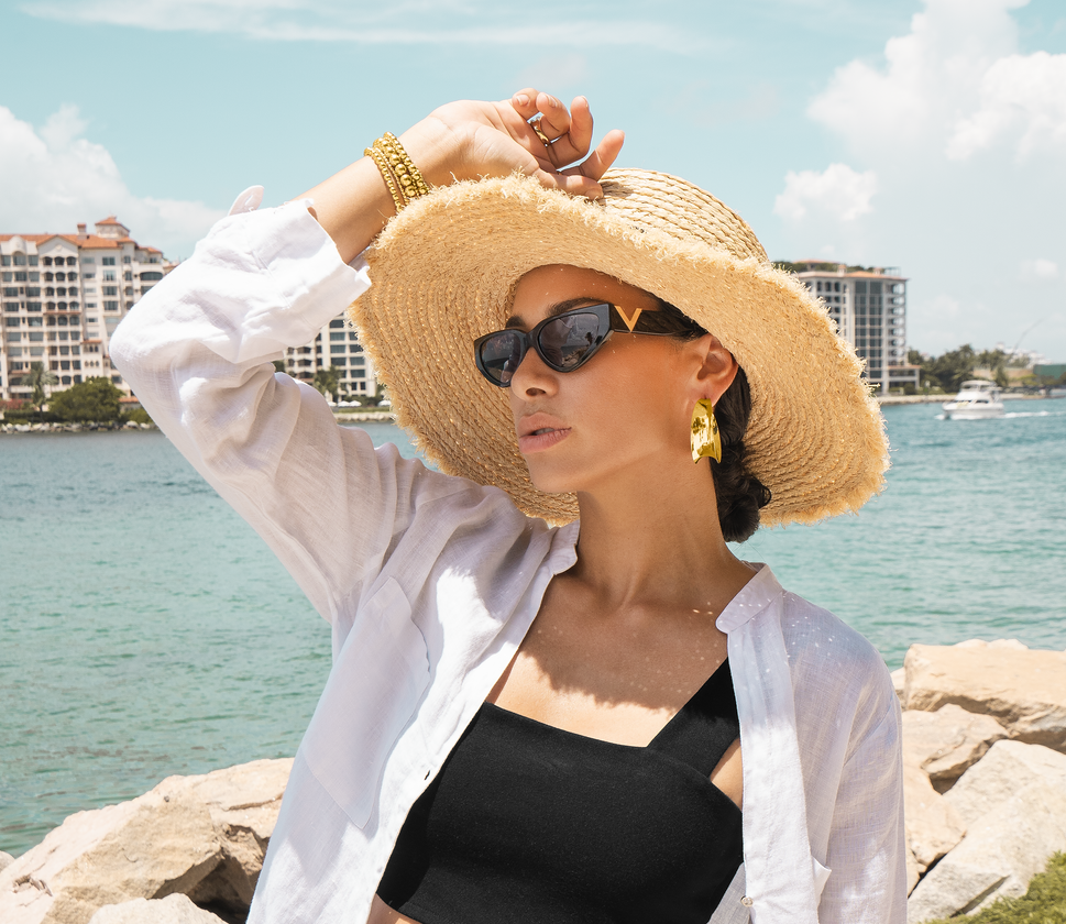 A stylish woman wearing chic cat-eye sunglasses and a wide-brimmed straw hat adjusts her sun hat by the water.