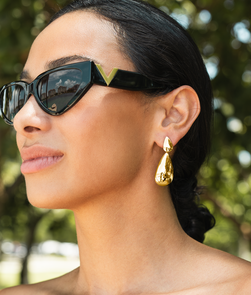 A stylish woman wearing chic black cat-eye sunglasses showcases a pair of elegant, polished gold teardrop earrings outdoors.