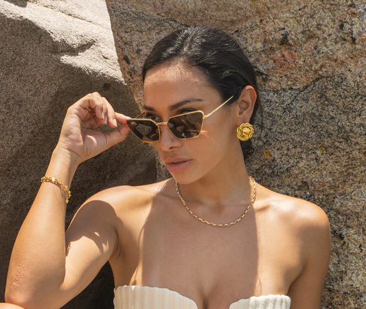 A glamorous woman wearing gold geometric sunglasses, a chunky gold chain necklace, and statement swirl earrings poses against a rocky background while adjusting her eyewear.