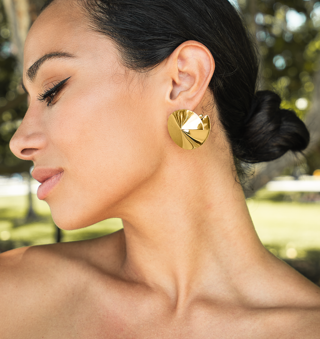 A woman wearing a large, textured gold statement earring showcases her profile against a softly blurred park background.