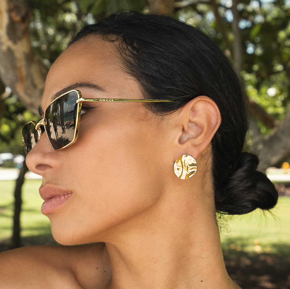 A woman wearing gold textured statement earrings and designer sunglasses poses outdoors in a park.