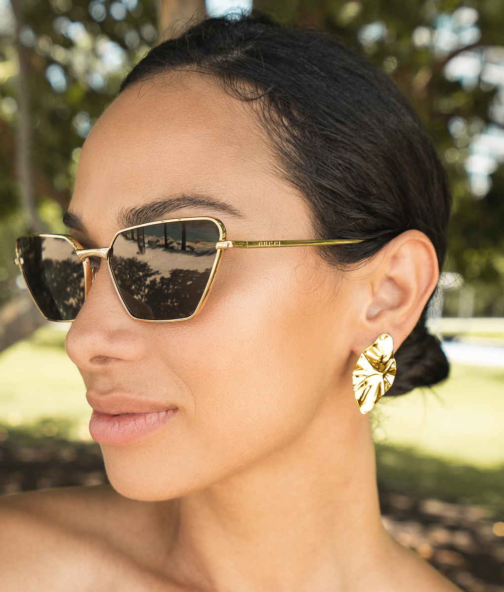 A woman wearing Gucci gold cat-eye sunglasses and statement leaf earrings poses outdoors.