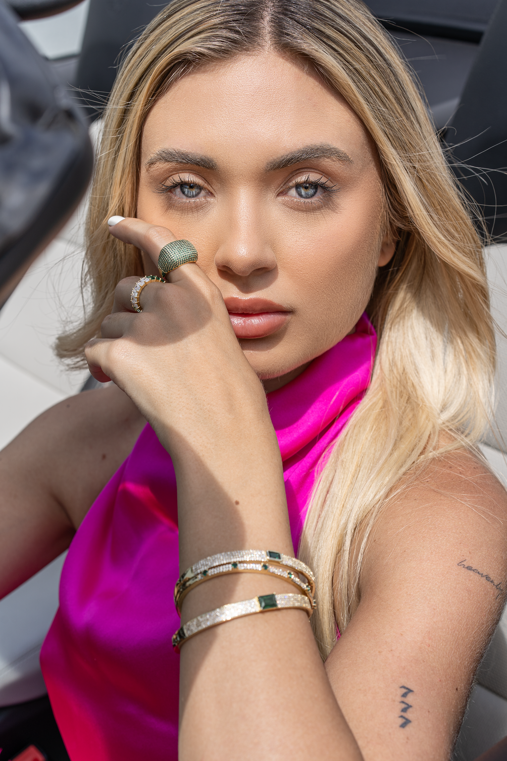 A blonde woman in a vibrant pink top showcases sparkling gold gemstone jewelry, including diamond bangles and a green pavé ring.