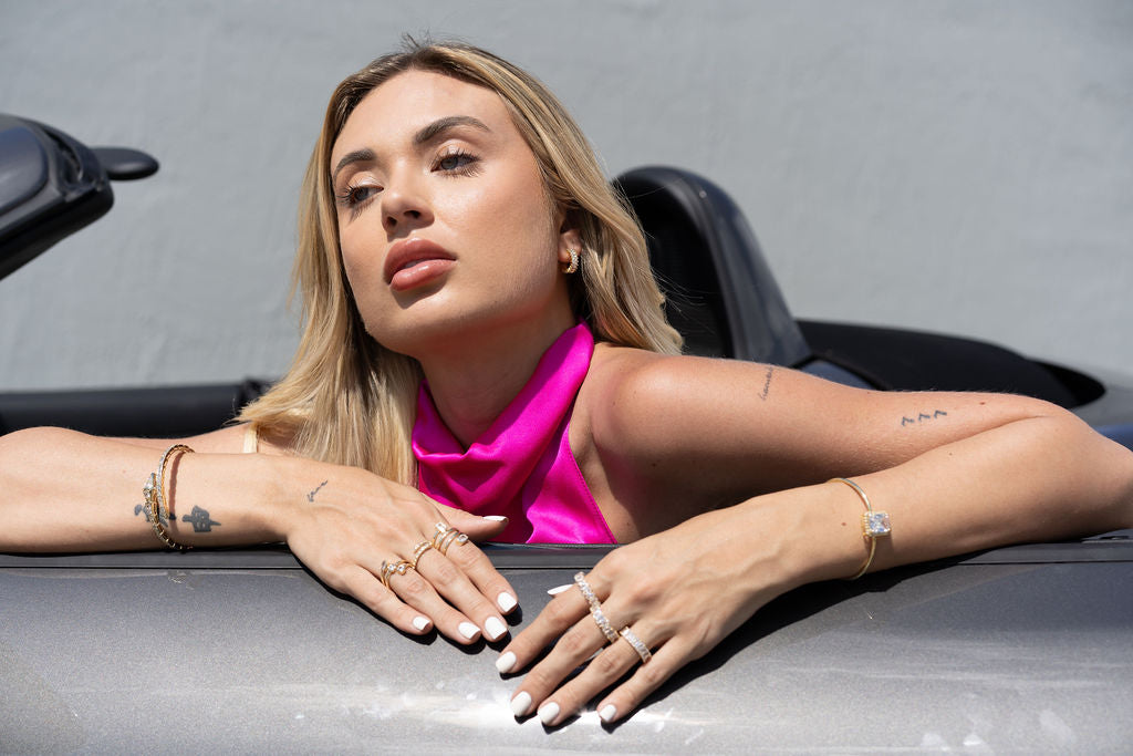 A blonde woman wearing a vibrant pink halter top and stacked gold jewelry poses in a convertible car.