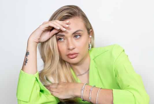 A blonde woman wearing a vibrant lime green blazer models sparkling diamond bracelets and earrings while resting her hand gently on her forehead.