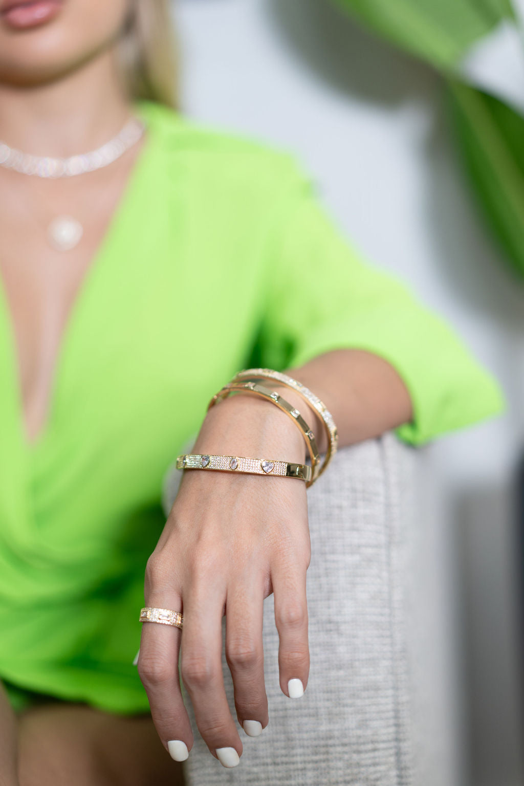 A woman wearing a vibrant green top showcases a stack of sparkling gold diamond bangles and a matching ring on her hand.