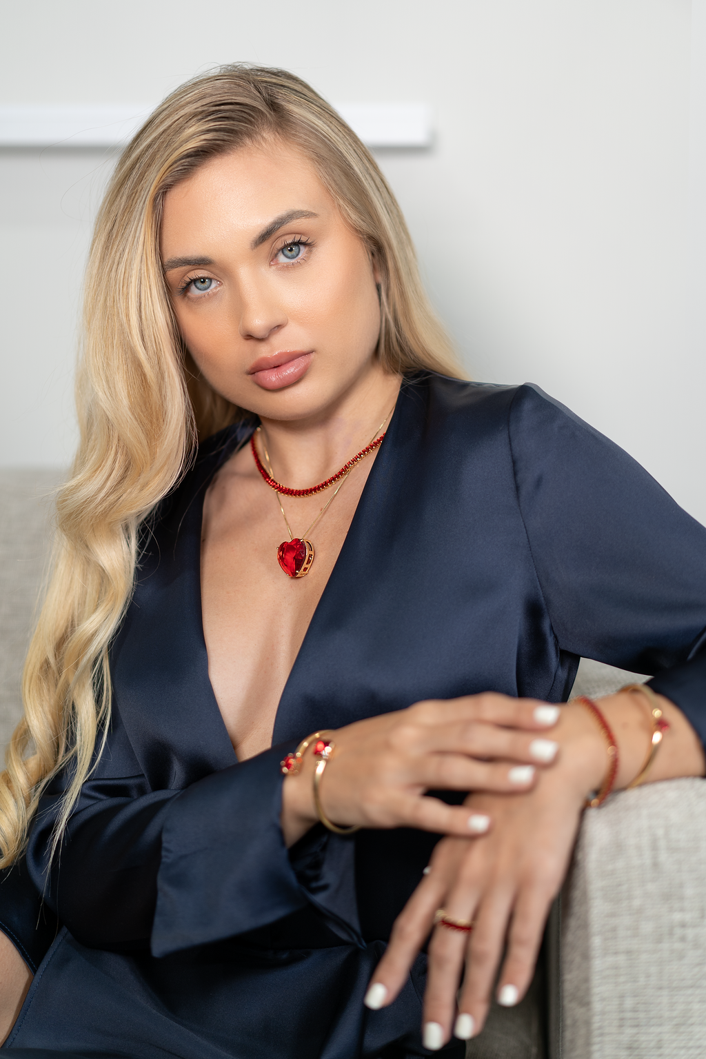 A blonde woman wearing a deep navy blue satin robe accessorizes with a stunning red heart pendant necklace and matching red crystal jewelry while posing gracefully on a sofa.
