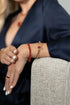 A woman wearing a gold ruby tennis bracelet and a gold bangle with a red gemstone rests her arm on a textured gray couch while wearing a navy silk robe.