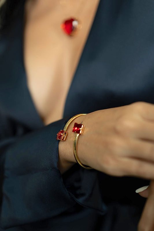 A woman wears a sleek gold open-cuff bracelet featuring square red gemstones on her wrist, complemented by a matching red heart pendant necklace against a dark silk blouse.