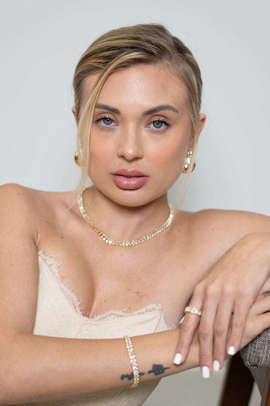 A blonde woman with blue eyes wears a sparkling tennis necklace, matching earrings, and a delicate bracelet while posing elegantly.