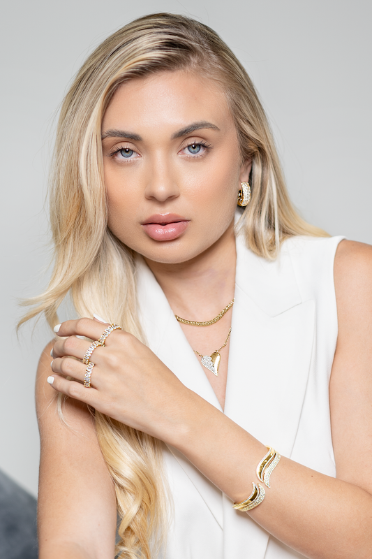 A blonde woman wearing sparkling gold jewelry, including layered necklaces, statement rings, and a cuff bracelet, poses gracefully against a neutral background.