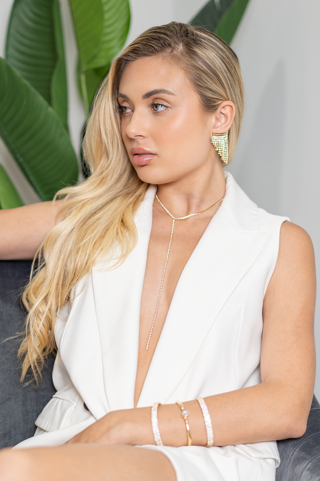 A stylish blonde model wearing a white vest showcases sparkling crystal fringe earrings, a delicate lariat necklace, and matching gemstone bracelets.